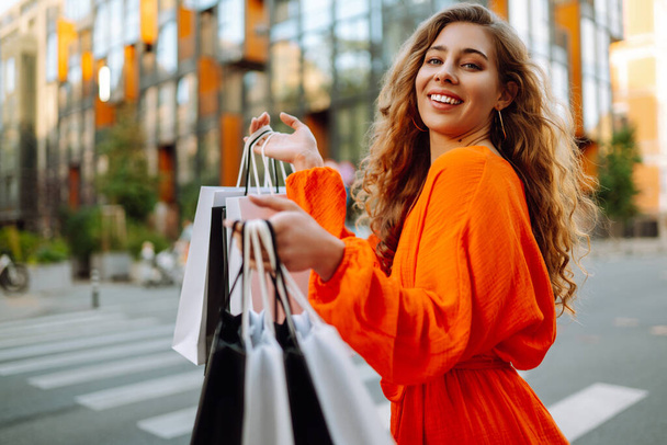Stylish woman with shopping bags walks through the city streets.  Consumerism, purchases, shopping, lifestyle concept. - 写真・画像