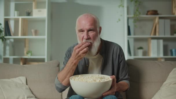 Een oude man die op de bank zit, popcorn eet en een enge film kijkt. Filmgenres en emotionele reactie. Online bioscopen. - Video