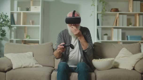 Een oude man in VR-bril speelt computerspellen met een Joystick, zit in de bank en eet een popcorn.Een moderne oudere man speelt PlayStation. Augmented Reality en Gaming voor senioren. - Video