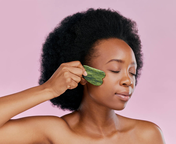 Gua sha, massage and black woman with natural beauty, face care and skin glow on pink background. Female model, jade cosmetic tools and dermatology with afro hair and facial skincare in studio. - Photo, Image