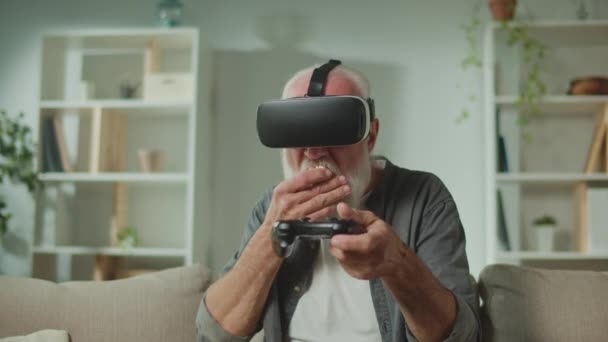 Un anciano en gafas VR juega juegos de ordenador con un joystick, sentado en el sofá y comiendo palomitas. Un anciano moderno juega PlayStation. Realidad Aumentada y Juego para Mayores. - Metraje, vídeo