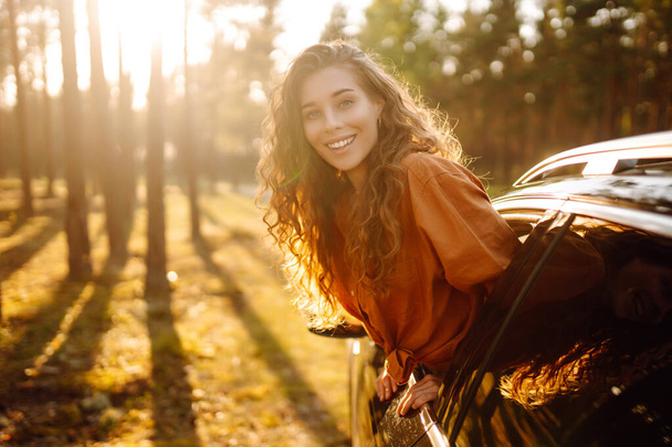 Beautiful traveler woman is resting and enjoying the sunset in the car. Happy woman on a summer trip, travels leaning out of the car window. Active lifestyle, tourism. Travel concept. - Фото, изображение