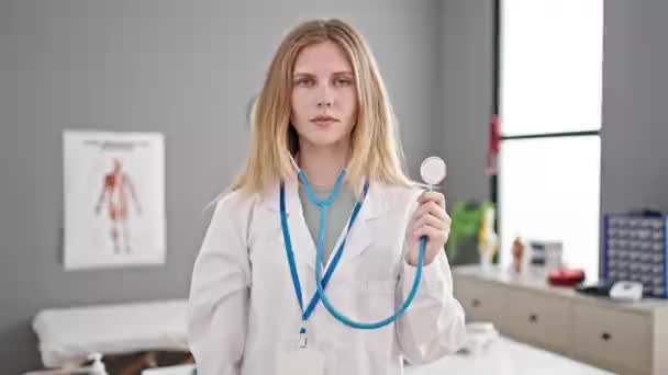 Νεαρή ξανθιά γυναίκα γιατρός χαμογελά αυτοπεποίθηση κρατώντας στηθοσκόπιο στην κλινική - Πλάνα, βίντεο