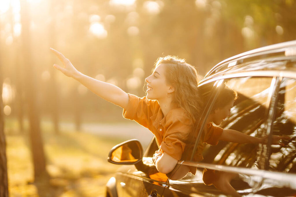 Beautiful traveler woman is resting and enjoying the sunset in the car. Happy woman on a summer trip, travels leaning out of the car window. Active lifestyle, tourism. Travel concept. - Foto, Imagen