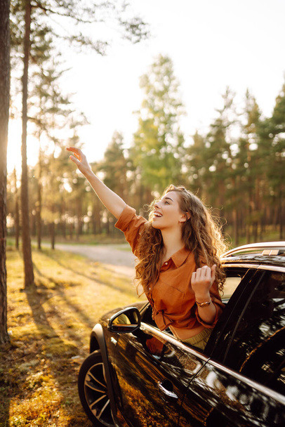 Beautiful traveler woman is resting and enjoying the sunset in the car. Happy woman on a summer trip, travels leaning out of the car window. Active lifestyle, tourism. Travel concept. - Foto, afbeelding