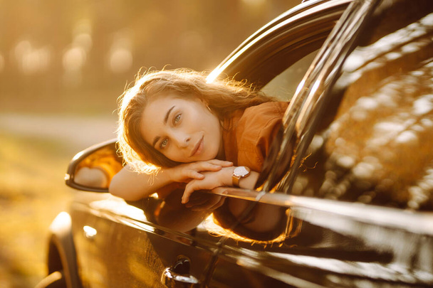 Belle voyageuse se repose et profite du coucher de soleil dans la voiture. Femme heureuse en voyage d'été, voyage penchée par la fenêtre de la voiture. Mode de vie actif, tourisme. Concept de voyage. - Photo, image