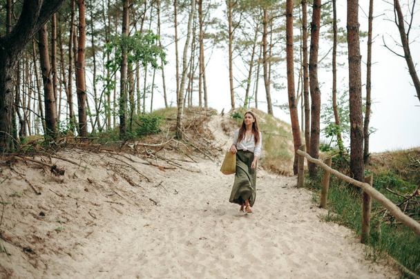 Front view of pretty young woman going down on sandy pathway and carrying big woven bag. Slender lady dressed in long dark-green skirt and white blouse enjoying forest landscape. - Foto, Imagem