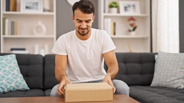 Joven hombre hispano desempacando caja de cartón sentado en el sofá con gesto ganador en casa - Imágenes, Vídeo