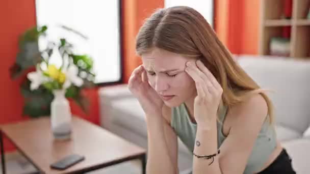 Νεαρή ξανθιά γυναίκα που υποφέρει από πονοκέφαλο κάθεται στον καναπέ στο σπίτι - Πλάνα, βίντεο