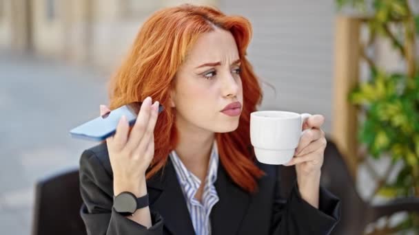 Giovane rossa donna d'affari udente messaggio vocale da smartphone bere caffè sulla terrazza della caffetteria - Filmati, video