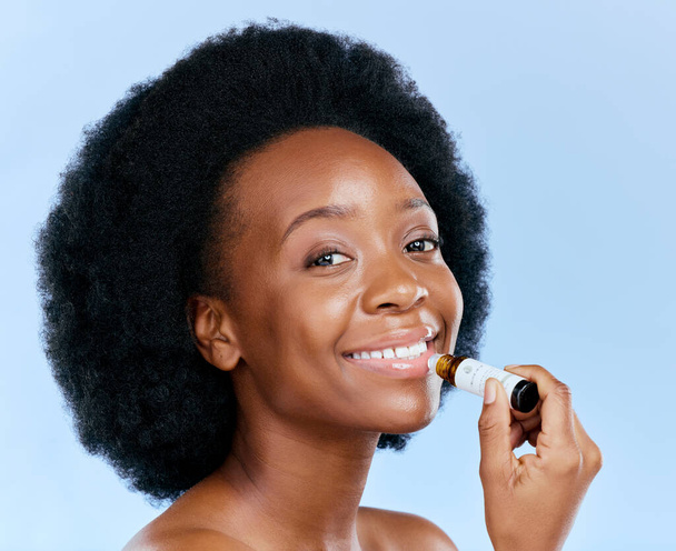Portrait, beauty and lip balm with a black woman in studio on a blue background for skincare cosmetics. Face, hydration and moisture with a happy young female model holding a makeup product for care. - Фото, изображение