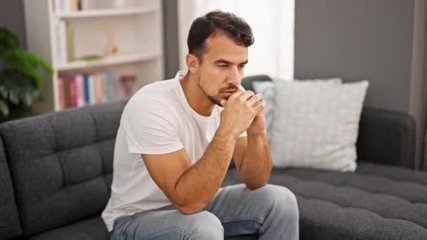Joven hombre hispano sentado en un sofá con expresión seria diciendo no en casa - Metraje, vídeo