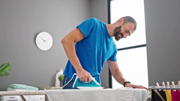 Junger hispanischer Mann lächelt selbstbewusst beim Bügeln von Kleidung in der Waschküche - Filmmaterial, Video