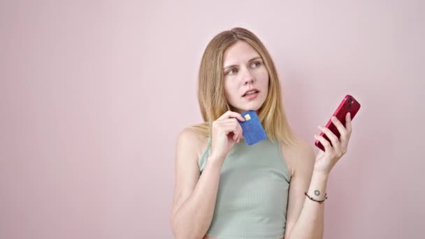 Nuori blondi nainen, jolla on älypuhelin ja luottokortti, jolla on idea eristetystä vaaleanpunaisesta taustasta - Materiaali, video