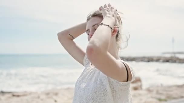 Nuori blondi nainen turisti hymyilee luottavainen koskettaa hiukset rannalla - Materiaali, video