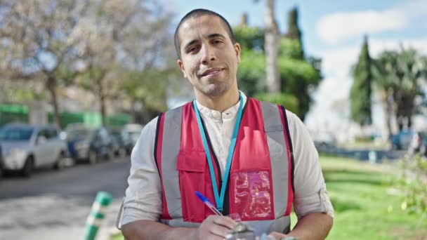 Jonge Spaanse man vrijwilliger glimlachend schrijven op klembord in park - Video