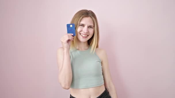 Молодая блондинка улыбается уверенно держа кредитку на изолированном розовом фоне - Кадры, видео