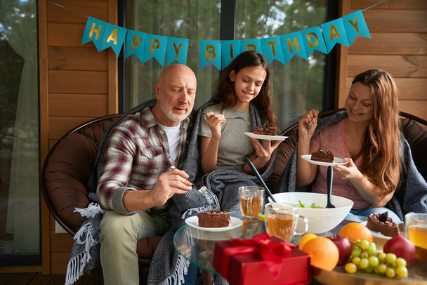 Fröhliches Mädchen und ihre Eltern sitzen mit Kuchenstücken auf dem Teller am Tisch auf der Veranda - Foto, Bild
