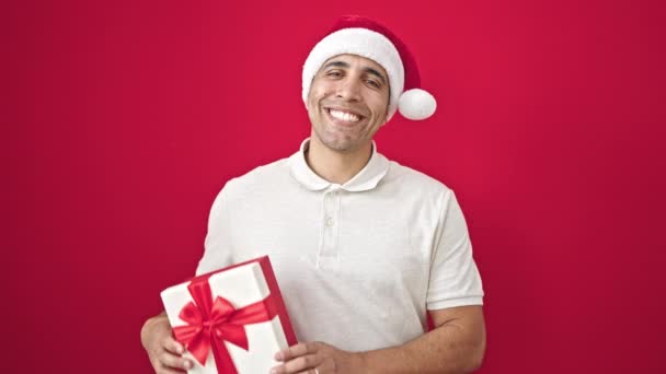Junger hispanischer Mann lächelt selbstbewusst und hält Weihnachtsgeschenk vor isoliertem roten Hintergrund - Filmmaterial, Video