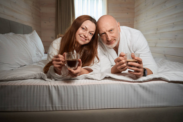 Усміхнена жінка і її чоловік лежать в зручному ліжку з чашками кави в руках - Фото, зображення