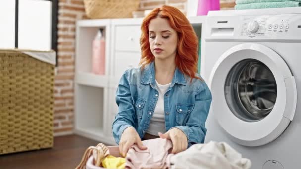 Junge rothaarige Frau sitzt auf dem Fußboden und faltet Kleidung in der Waschküche - Filmmaterial, Video