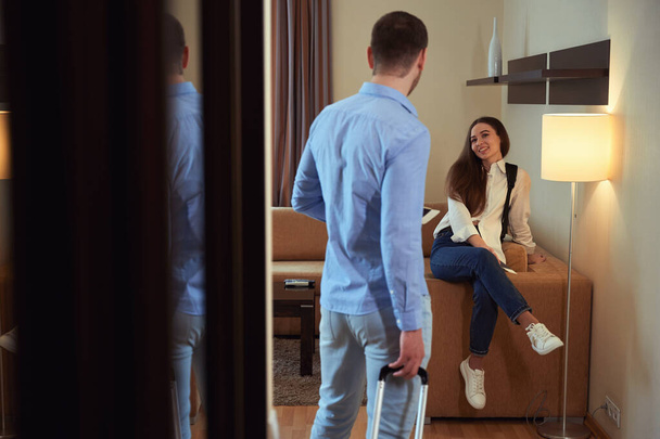 El hombre mira a su novia sentada en un sofá en una habitación de hotel. Vista desde atrás - Foto, Imagen