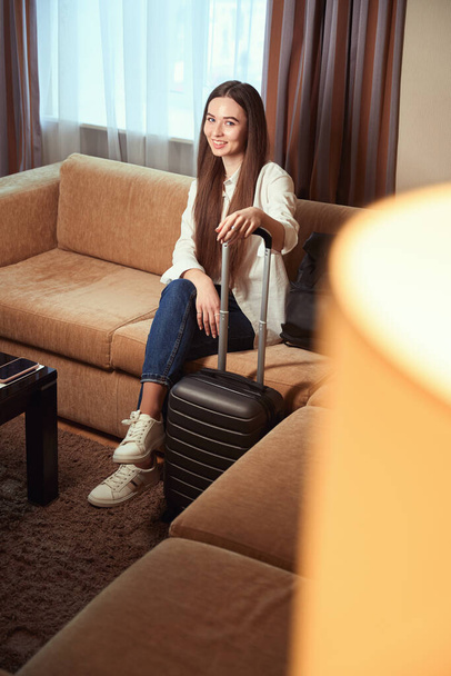 Portrait of smiling woman in hotel room on sofa with luggage nearby. Check in hotel - Foto, Bild