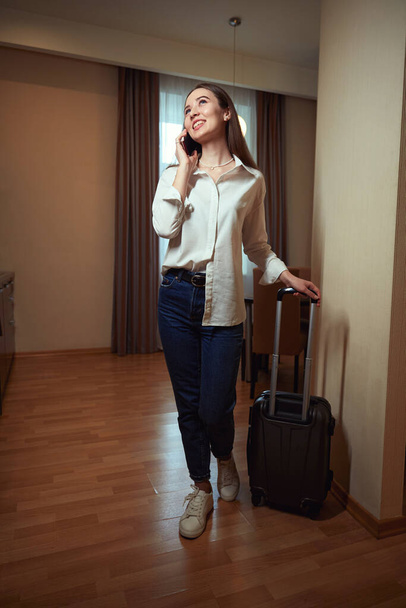 Happy lady talking on phone and holding suitcase by handle in hotel room - Photo, Image