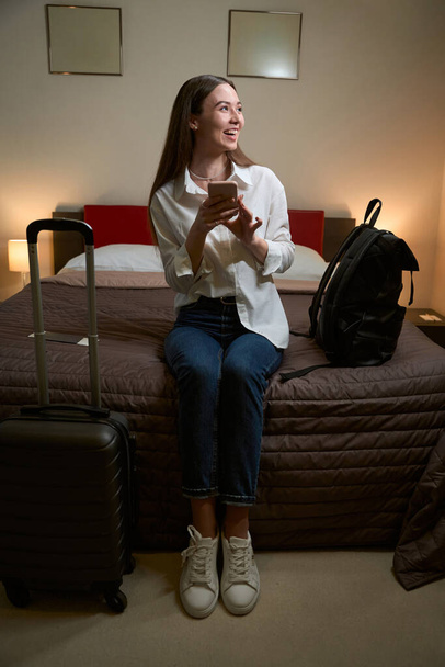 Piękna pani trzyma telefon i siedzi na kanapie z walizką obok niej w swoim pokoju. Zameldowanie w hotelu - Zdjęcie, obraz