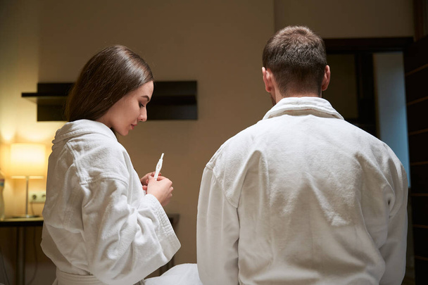 Young woman seated on bed beside man in bathrobe looking at pregnancy test device in her hands - Photo, Image