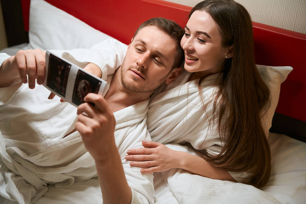 Женщина лежит в постели в номере отеля, показывая супругу ультразвуковые снимки первого триместра - Фото, изображение