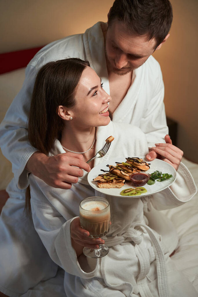 Romantische man trakteert zijn vrolijke vrouwelijke metgezel met pannenkoeken in hotelsuite - Foto, afbeelding