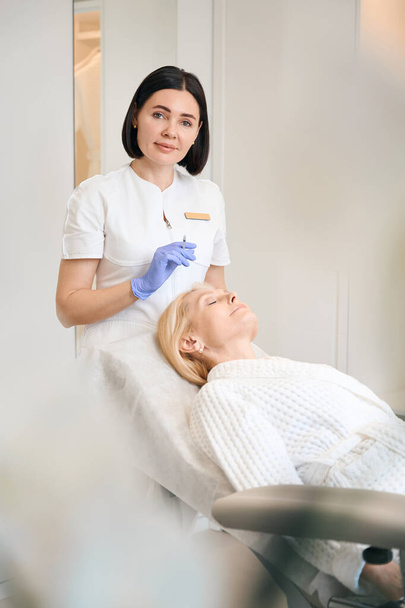 Happy cosmetologist holding syringe above mature woman face in beauty salon - Photo, Image