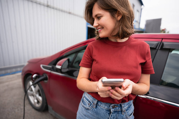 Woman with smartphone charging red electric car - Φωτογραφία, εικόνα
