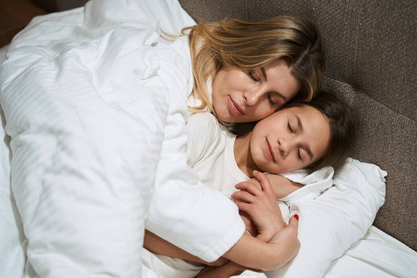 Mãe deitada na cama perto da menina e abraçando-a no quarto do hotel - Foto, Imagem