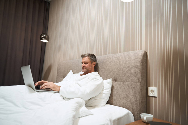 Adult man in white coat resting in bed and typing on laptop in the hotel - Φωτογραφία, εικόνα