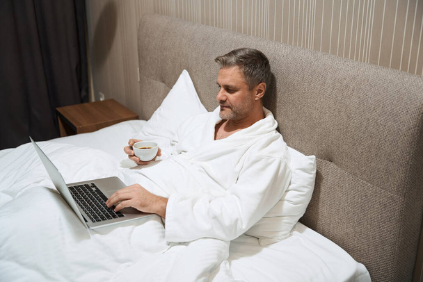 Mann in weißem Mantel liegt auf Bett und tippt am Computer, hält Tasse im Motel - Foto, Bild