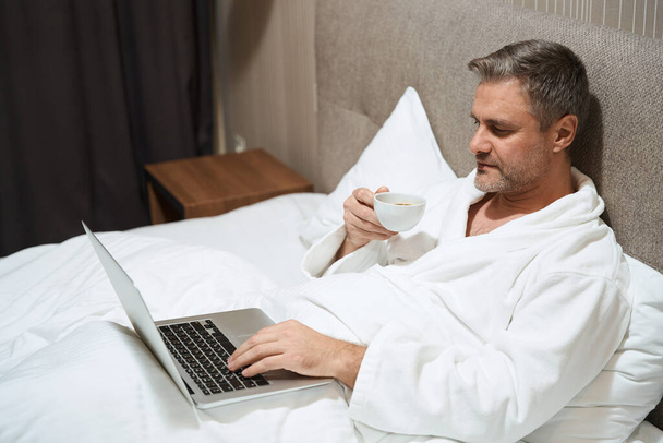 Adult male in white coat resting on bed and typing on laptop, drinking coffee in the hotel - 写真・画像