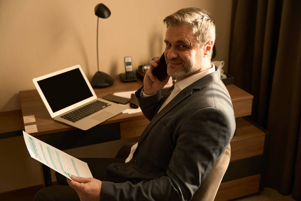 Adult man in business suit sitting near laptop and calling on smartphone, holding papers in the hotel room - Foto, Bild