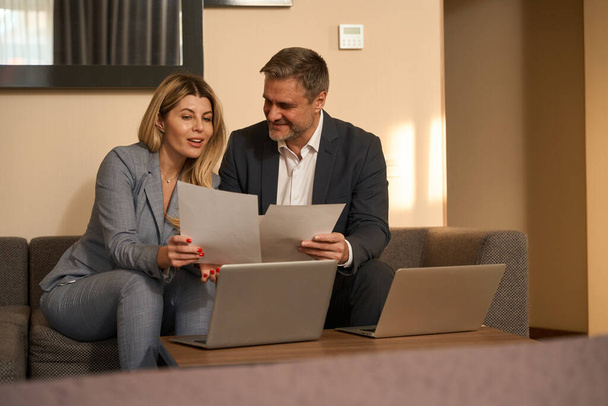 Adult man and smiling woman in business suits sitting on sofa and working, discussion documents in the motel - Fotó, kép