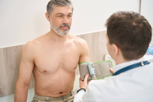 Lääkäri valkoisessa takissa istuu lähellä miestä ja mittaa painetta klinikalla - Valokuva, kuva
