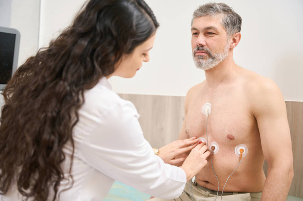 Verpleegster in witte vacht voorbereiding patiënt voor elektrocardiografie van het hart, man op zoek naar arts - Foto, afbeelding