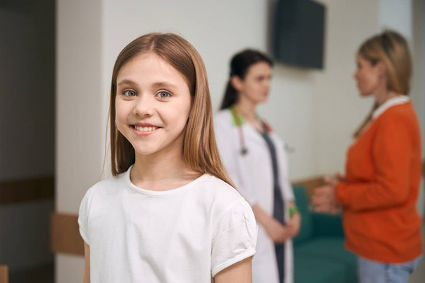 Happy girl patient of pediatrician clinic looking at camera and smiling, while mother discussing her health test results with doctor - Фото, зображення