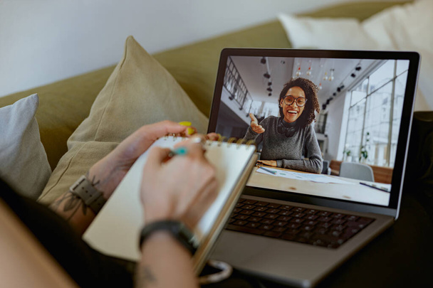 Woman using laptop for online learning and making notes while lying on sofa at home - Photo, Image
