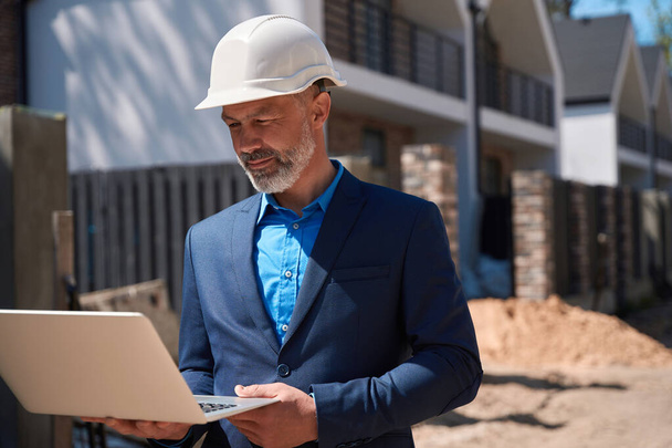 Геодезист-мужчина в деловом костюме и каскаде осматривает строительную площадку, осматривает землю с ноутбуком в руках, исследует ландшафт - Фото, изображение