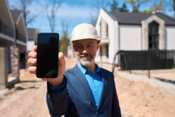 Smiling foreman in hardhat holding and showing smartphone display, showing 3d model of ready townhouse standing at construction site - Fotoğraf, Görsel