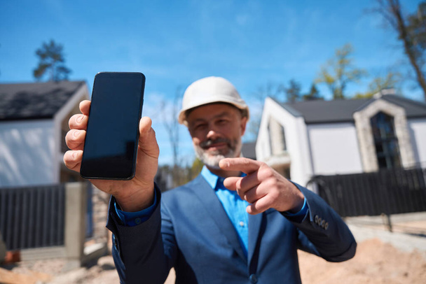 Smiling man building manager in hardhat pointing finger at smartphone display standing at construction site, sale and rental of residential buildings - Valokuva, kuva