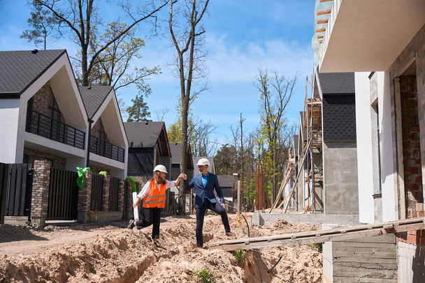 Foreman in business suit and hardhat showing to female investor her future house, walking along construction site, inspectors checking compliance of safety regulations - Photo, image