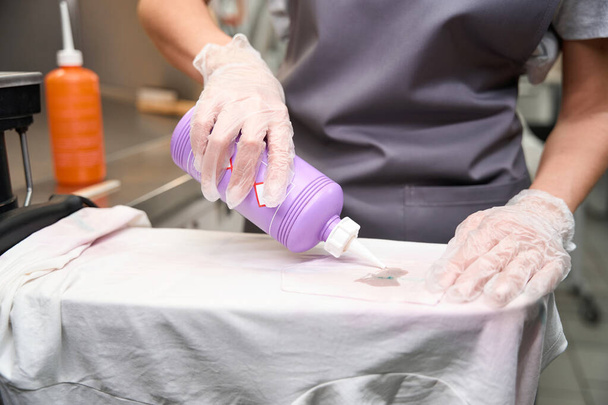 Woman in protective gloves applying solvent into big spot on white t-shirt, professional clothing cleaning, dry-cleaning service - Valokuva, kuva