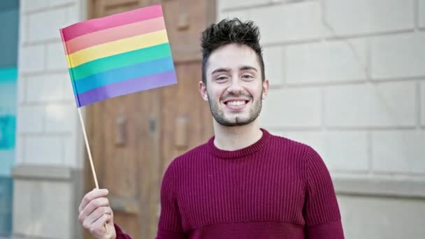 Fiatal spanyol férfi mosolyog magabiztosan tartva szivárvány zászló az utcán - Felvétel, videó